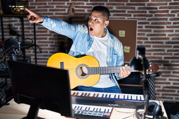 Hispanic young man playing classic guitar at music studio pointing with finger surprised ahead, open mouth amazed expression, something on the front