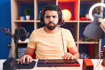 Hispanic man with beard playing video games with headphones puffing cheeks with funny face. mouth inflated with air, crazy expression.