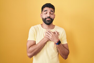 Hispanic man with beard standing over yellow background smiling with hands on chest with closed eyes and grateful gesture on face. health concept.