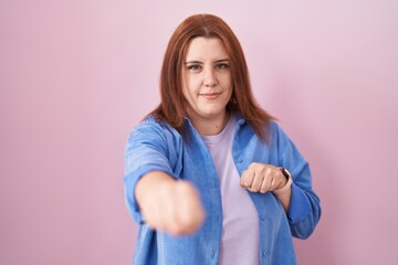 Young hispanic woman with red hair standing over pink background punching fist to fight, aggressive and angry attack, threat and violence