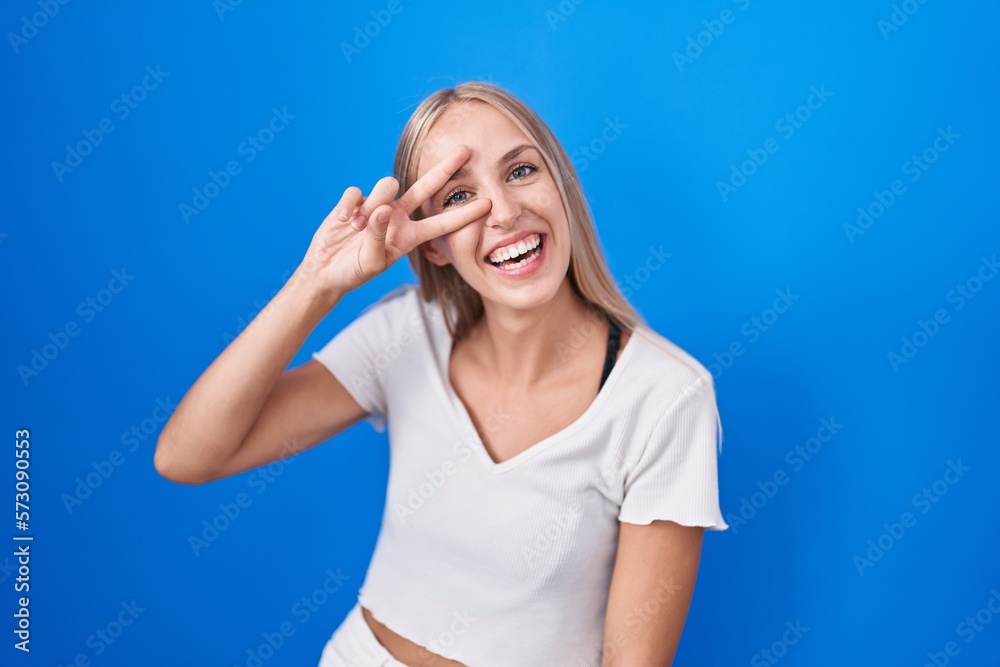 Sticker Young caucasian woman standing over blue background doing peace symbol with fingers over face, smiling cheerful showing victory