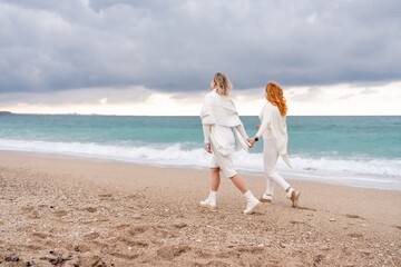 Fototapeta na wymiar Women sea walk friendship spring. Two girlfriends, redhead and blonde, middle-aged walk along the sandy beach of the sea, dressed in white clothes. Against the backdrop of a cloudy sky and the winter