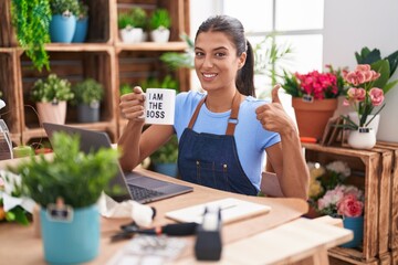 Brunette young woman working at florist shop holding i am the boss cup smiling happy and positive, thumb up doing excellent and approval sign