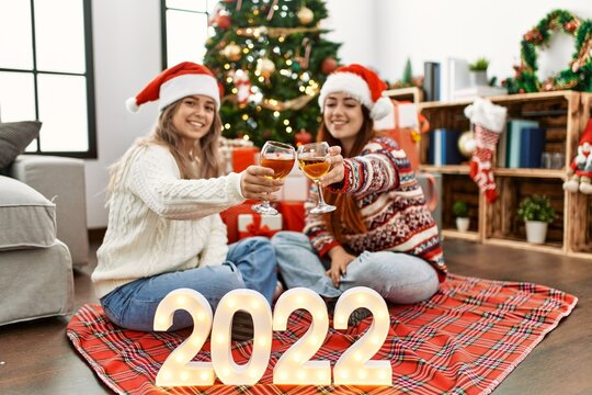 Woman couple toasting with champagne for new year 2022 sitting by christmas tree at home