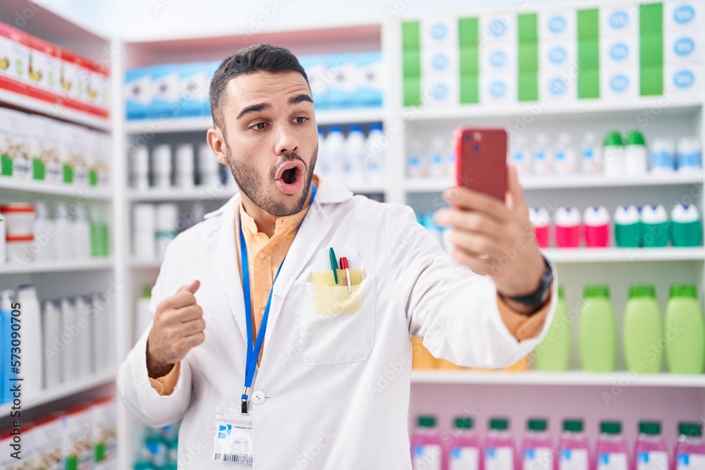 Sticker young hispanic man working at pharmacy drugstore doing video call with smartphone scared and amazed 