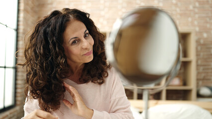Middle age hispanic woman combing hair with hand sitting on bed at bedroom