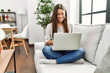 Young latin woman using laptop sitting on sofa at home - obrazy, fototapety, plakaty