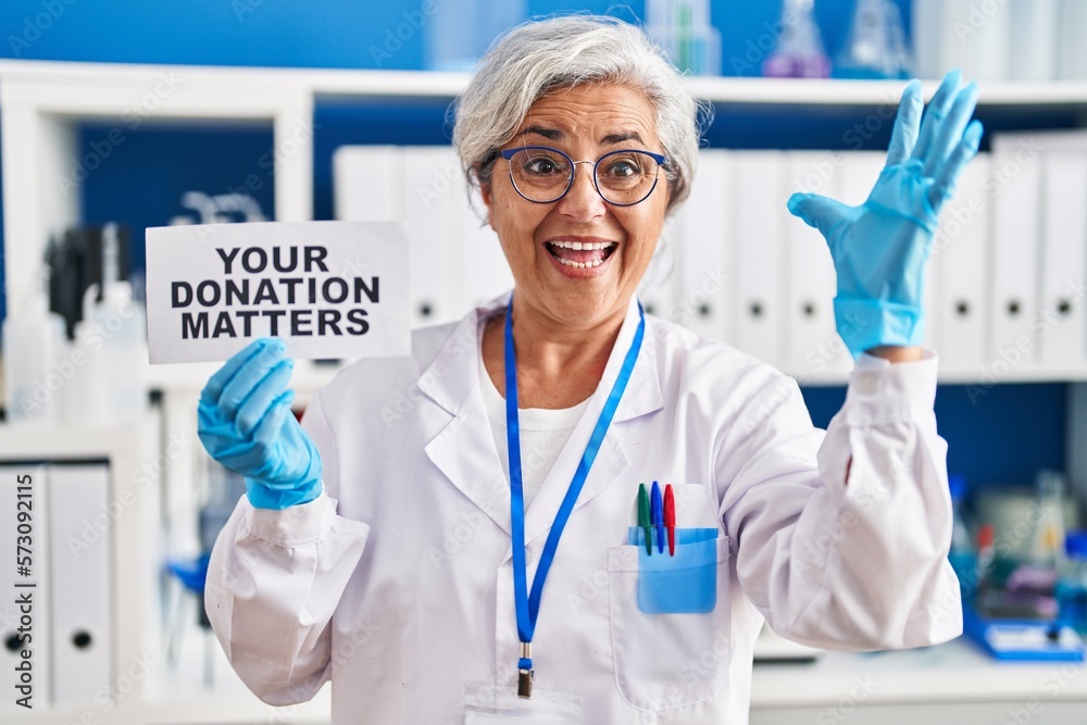 Poster Middle age woman with grey hair working at scientist laboratory holding your donation matters banner celebrating victory with happy smile and winner expression with raised hands