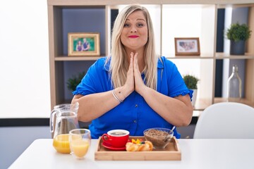 Obraz na płótnie Canvas Caucasian plus size woman eating breakfast at home praying with hands together asking for forgiveness smiling confident.