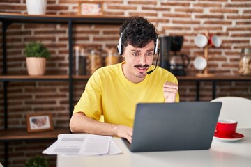 Hispanic man wearing call center agent headset working from home screaming proud, celebrating victory and success very excited with raised arms