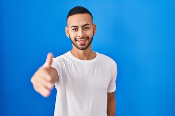 Young hispanic man standing over blue background smiling cheerful offering palm hand giving assistance and acceptance.