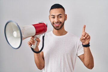 Young hispanic man shouting through megaphone smiling with an idea or question pointing finger with happy face, number one
