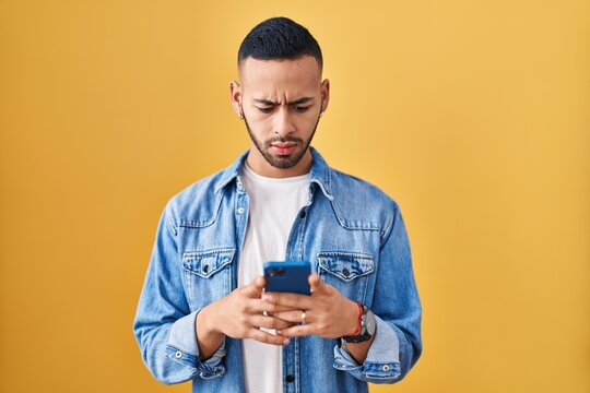 Young hispanic man using smartphone typing message skeptic and nervous, frowning upset because of problem. negative person.