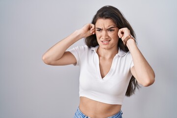 Young teenager girl standing over white background covering ears with fingers with annoyed expression for the noise of loud music. deaf concept.