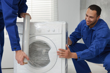 Obraz premium Male movers with stretch film wrapping washing machine in bathroom. New house