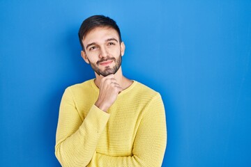 Hispanic man standing over blue background with hand on chin thinking about question, pensive expression. smiling and thoughtful face. doubt concept.