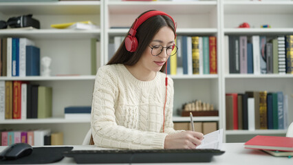 Young beautiful hispanic woman student using computer and headphones writing on notebook at library university