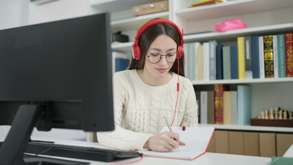Young beautiful hispanic woman student using computer and headphones writing on notebook at library university