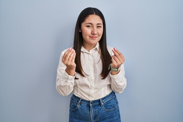 Young latin woman standing over blue background doing money gesture with hands, asking for salary payment, millionaire business