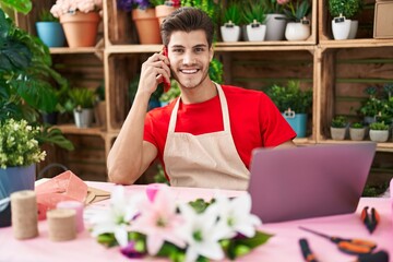 Young hispanic man florist talking on smartphone using laptop at flower shop