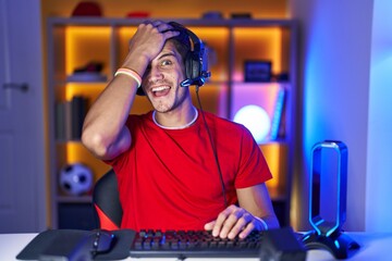 Young hispanic man playing video games surprised with hand on head for mistake, remember error. forgot, bad memory concept.