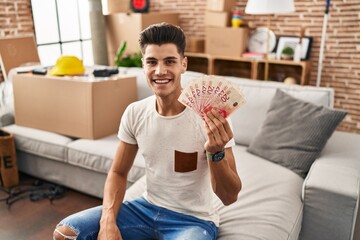 Young hispanic man moving to a new home holding shekels looking positive and happy standing and smiling with a confident smile showing teeth