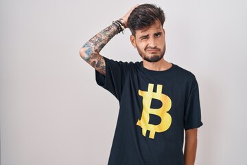 Young hispanic man with tattoos wearing bitcoin t shirt confuse and wondering about question....