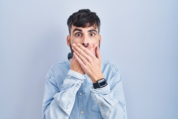 Young hispanic man with beard standing over blue background shocked covering mouth with hands for mistake. secret concept.
