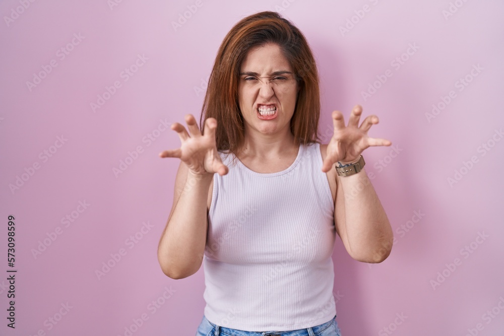 Poster brunette woman standing over pink background smiling funny doing claw gesture as cat, aggressive and