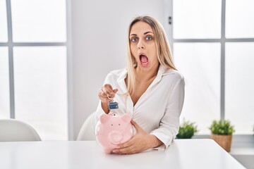 Young blonde woman holding piggy bank and house keys afraid and shocked with surprise and amazed expression, fear and excited face.