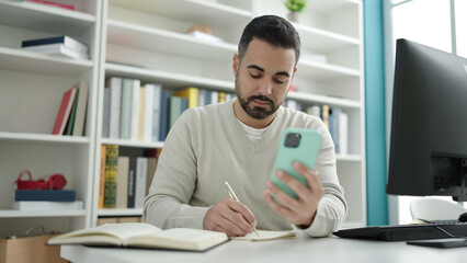 Young hispanic man student using smartphone writing on notebook at library university