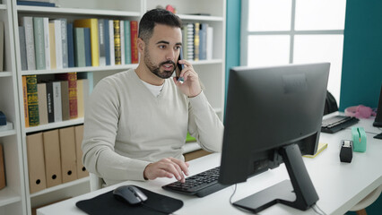 Young hispanic man student using computer talking on smartphone at library university