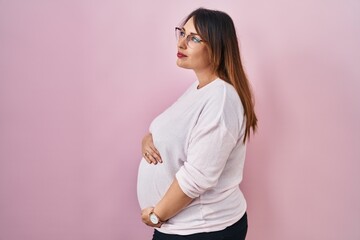 Pregnant woman standing over pink background looking to side, relax profile pose with natural face...