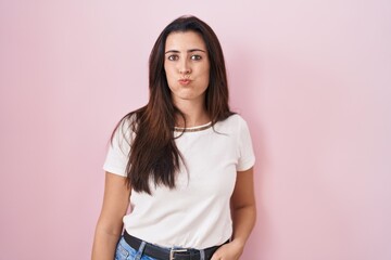 Young brunette woman standing over pink background puffing cheeks with funny face. mouth inflated with air, crazy expression.