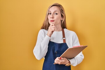 Young blonde woman wearing professional waitress apron holding clipboard serious face thinking...