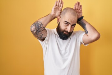 Young hispanic man with beard and tattoos standing over yellow background doing bunny ears gesture with hands palms looking cynical and skeptical. easter rabbit concept.