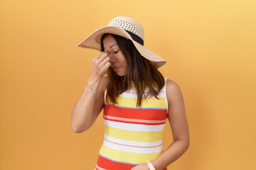 Middle age chinese woman wearing summer hat over yellow background tired rubbing nose and eyes feeling fatigue and headache. stress and frustration concept.