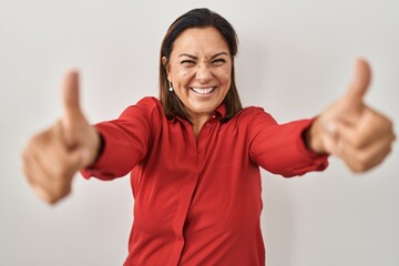 Hispanic mature woman standing over white background approving doing positive gesture with hand, thumbs up smiling and happy for success. winner gesture.