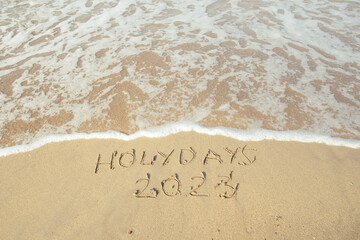Fototapeta premium Holidays 2023 lettering on the beach with wave and clear blue sea. 