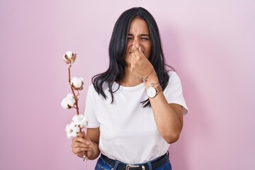 Brunette woman standing over pink background smelling something stinky and disgusting, intolerable...