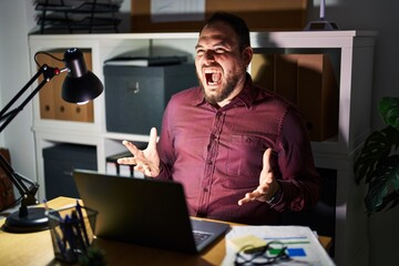Plus size hispanic man with beard working at the office at night crazy and mad shouting and yelling with aggressive expression and arms raised. frustration concept.
