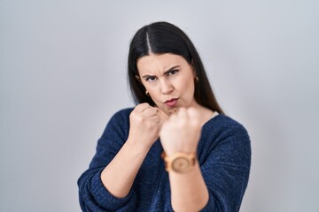 Young brunette woman standing over isolated background ready to fight with fist defense gesture, angry and upset face, afraid of problem