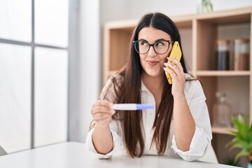 Young brunette woman holding pregnancy test result speaking on the phone smiling looking to the side and staring away thinking.