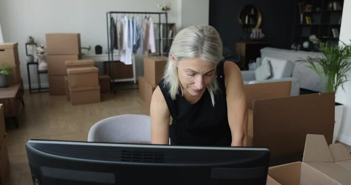 Woman typing on PC on relocation day, small business owner use computer, send e-mail to client working from home office with stacked carton boxes in living room like warehouse with goods for selling