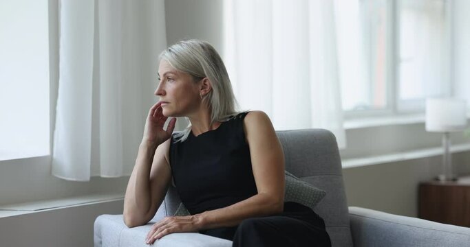 Middle age thoughtful, pensive woman sits on armchair staring out of window, ponders, thinking, missing or waiting, looks frustrated, recall life and recollects memories, resting alone in living room