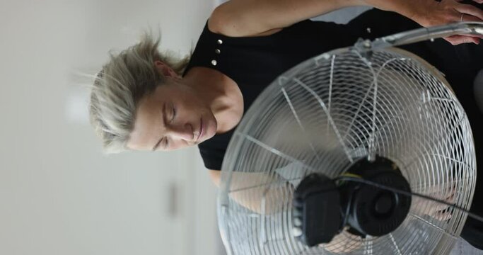 Middle-aged woman exhausted by unbearable heat, cooling herself using electric floor fan sit on sofa in living room. Extreme hot weather, need air-condition, climate control system, hormonal imbalance