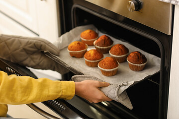 Young woman taking tray with cupcakes from oven in kitchen, closeup