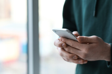 Young man with modern mobile phone near window, closeup