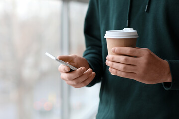 Young man with cup of coffee and modern mobile phone near window, closeup