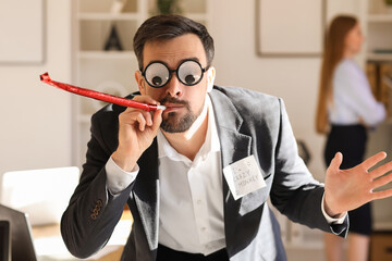Young man with funny eyeglasses and party blower in office. April Fools' Day celebration
